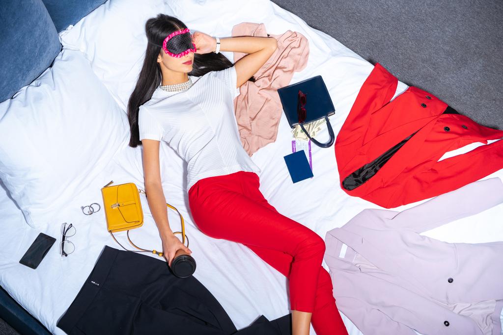 vista superior de la muchacha en máscara de ojos sosteniendo café para ir y dormir en la cama con ropa, pasaporte y dinero
 - Foto, Imagen
