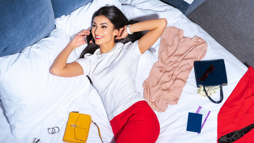 mooie lachende vrouw praten door smartphone terwijl liggend op bed met stijlvolle kleding, paspoort en instapkaart - Foto, afbeelding