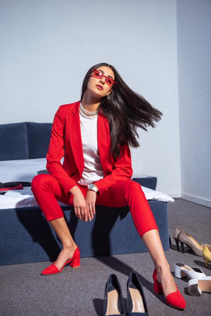 beautiful young woman in fashionable red suit and sunglasses sitting on bed  - Photo, Image