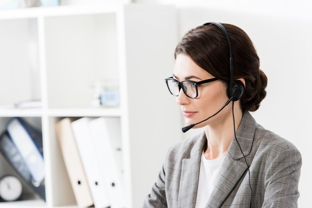 operador atractivo centro de llamadas en gafas y auriculares mirando hacia otro lado en la oficina
 - Foto, Imagen