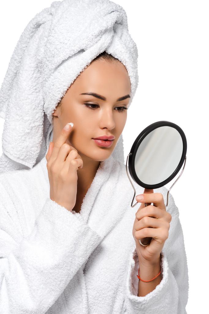 hermosa chica en albornoz mirando el espejo y la aplicación de crema en la cara aislada en blanco
 - Foto, imagen