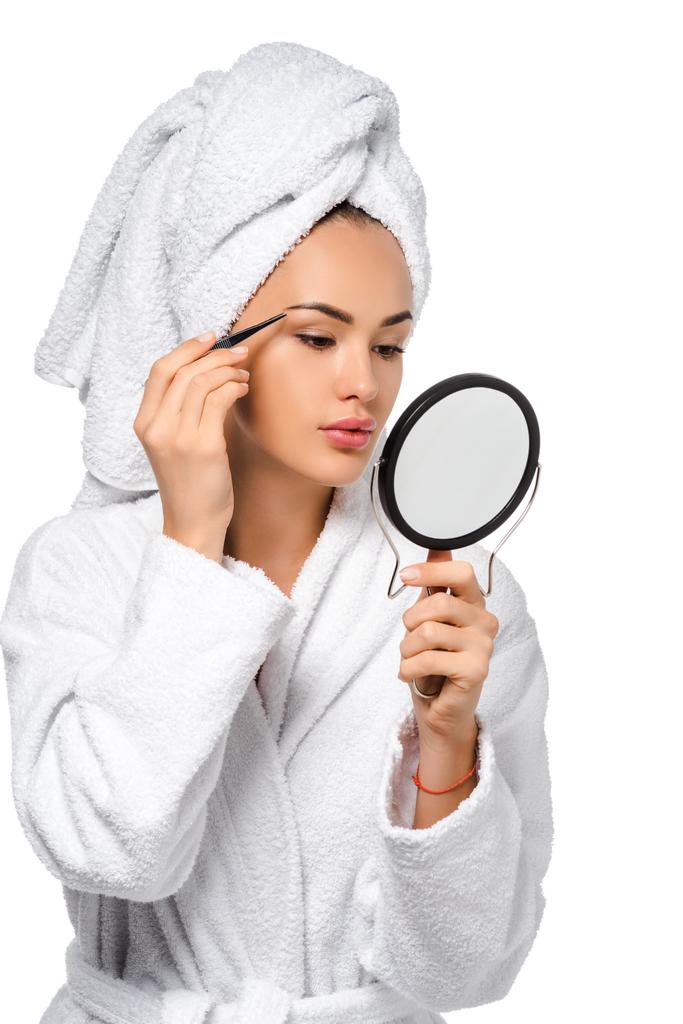 schöne Mädchen im Bademantel mit Blick auf Spiegel und Pinzette Augenbrauen isoliert auf weiß - Foto, Bild