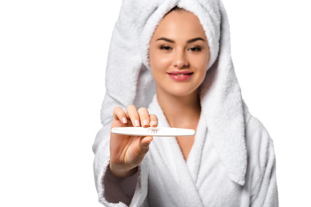 attraktives Mädchen im Bademantel lächelt, zeigt Schwangerschaftstest und blickt isoliert auf weiße Kamera - Foto, Bild