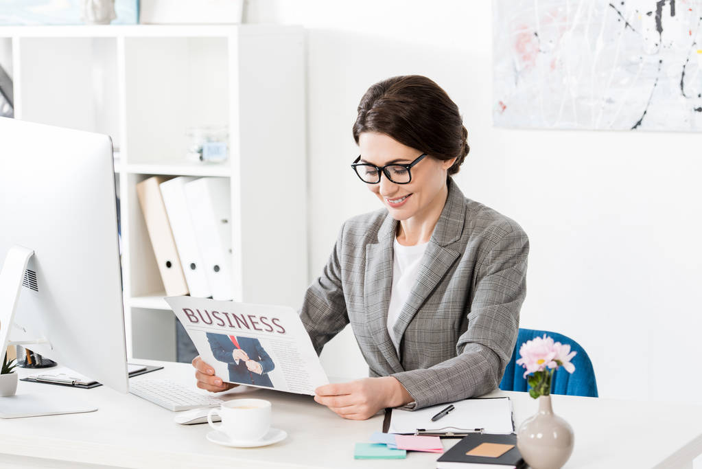 sonriente atractiva mujer de negocios en gafas leyendo periódico en la oficina
 - Foto, Imagen