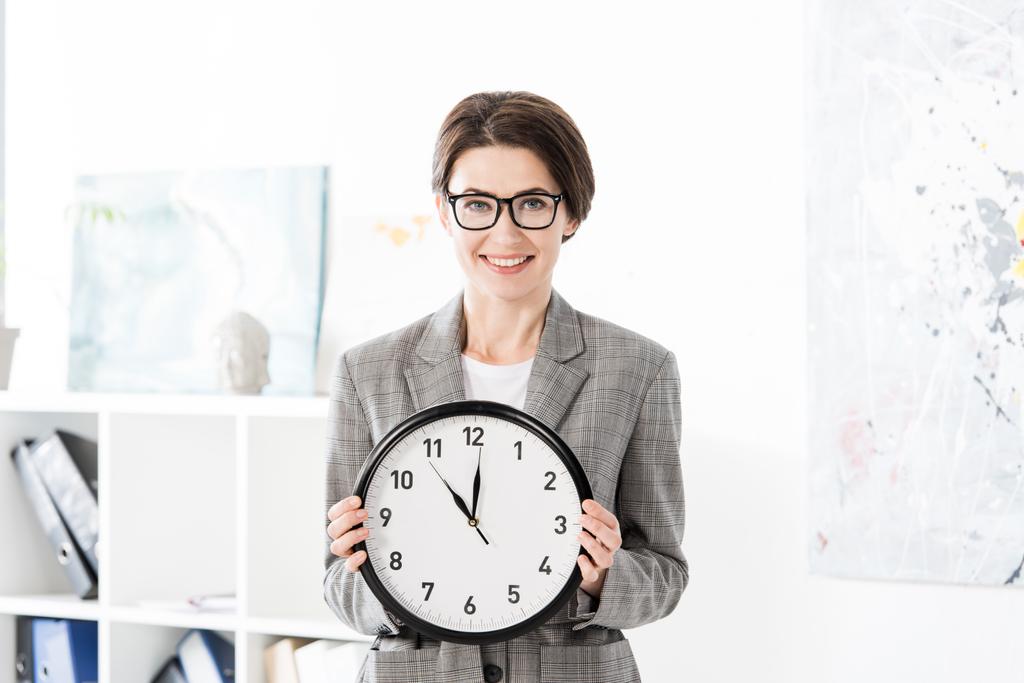 portrait of smiling attractive businesswoman holding wall clock and looking at camera in office - Photo, Image