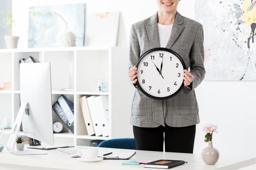 imagem cortada de mulher de negócios atraente segurando relógio de parede no escritório
 - Foto, Imagem