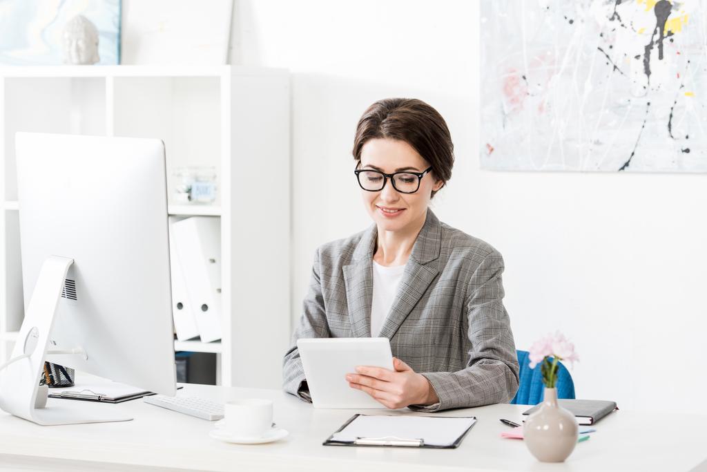 joyeuse femme d'affaires attrayante en costume gris en utilisant une tablette à la table au bureau
 - Photo, image