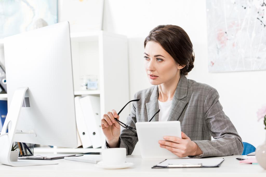 hermosa mujer de negocios en traje gris sosteniendo tableta y mirando a la computadora en la oficina
 - Foto, imagen