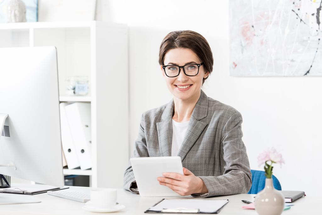 sonriente atractiva mujer de negocios en traje gris usando tableta y mirando a la cámara en la oficina
 - Foto, imagen