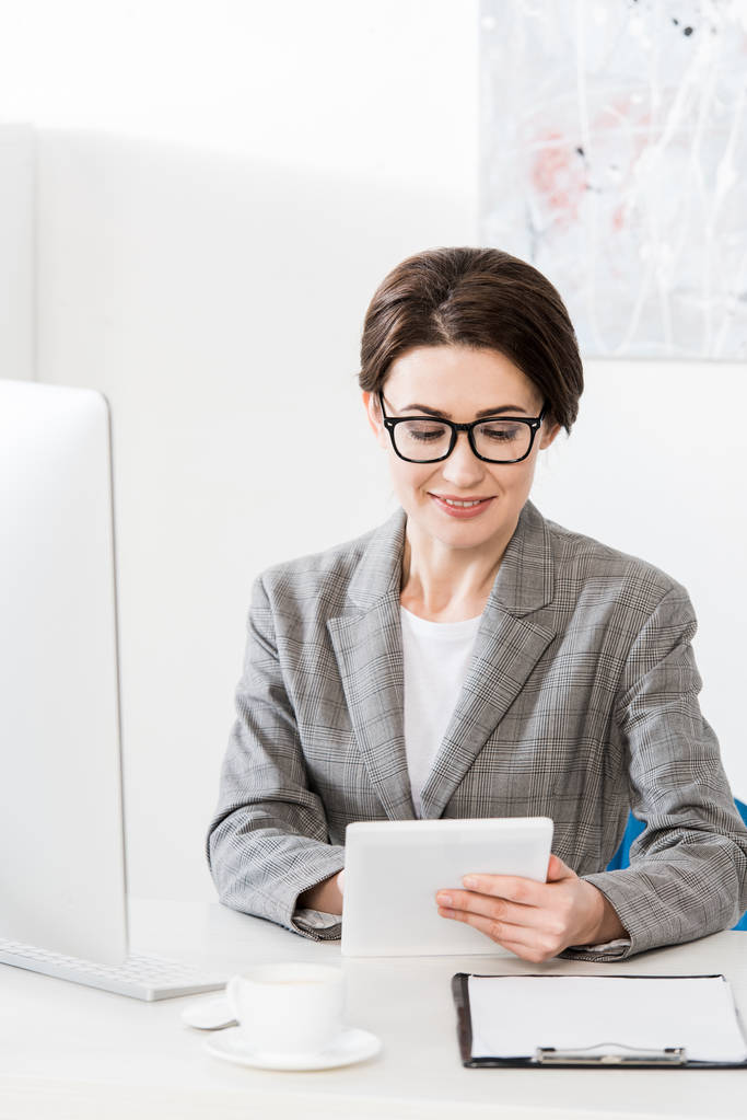 vrolijke aantrekkelijke zakenvrouw in grijs pak tablet gebruiken in office - Foto, afbeelding