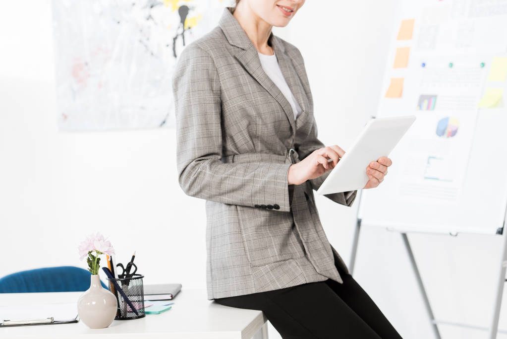 cropped image of businesswoman in grey suit sitting on table and using tablet in office - Photo, Image