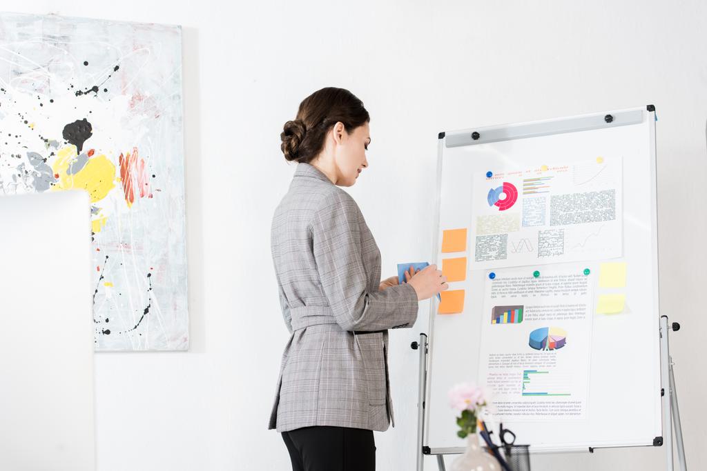 vista lateral de empresária atraente em terno cinza em pé perto flipchart no escritório
 - Foto, Imagem