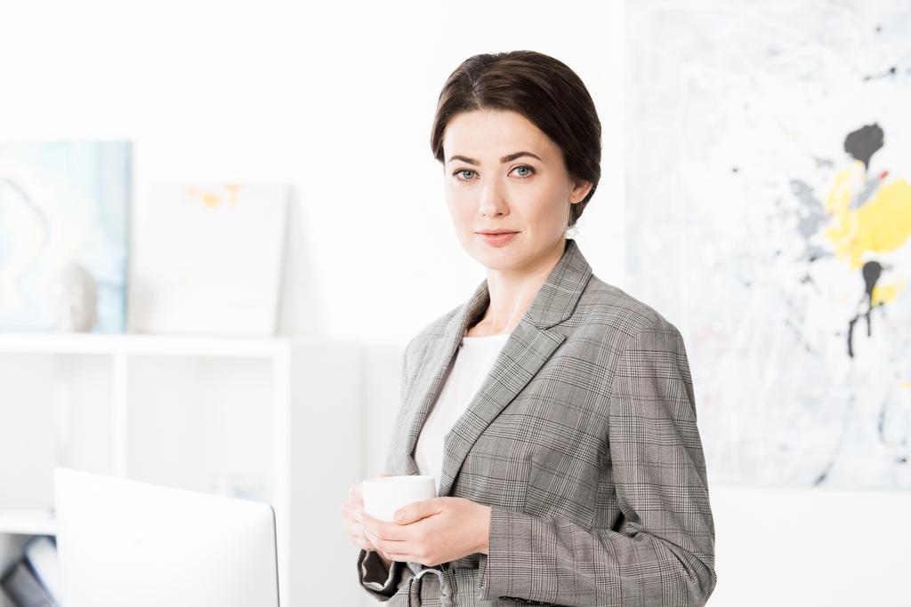 atractiva mujer de negocios en traje gris sosteniendo taza de té y mirando a la cámara en la oficina
 - Foto, imagen