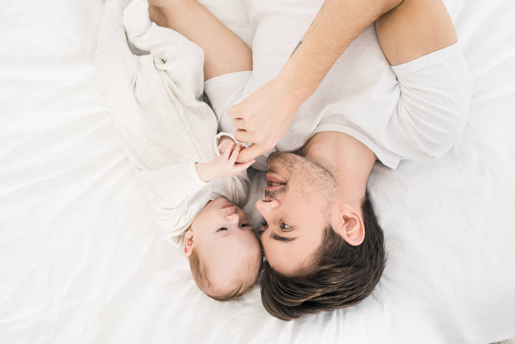 若い父親と家庭で一緒にベッドの上に横たわる小さな赤ちゃんのオーバー ヘッド ビュー - 写真・画像