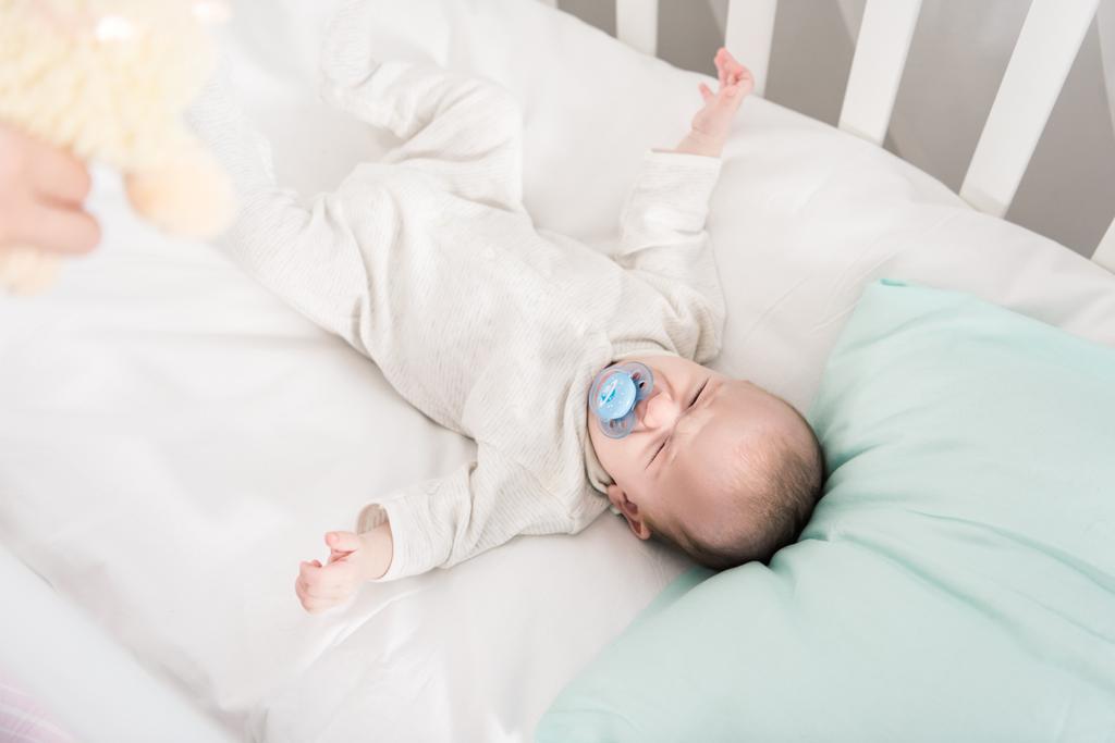 частичный вид родителя, показывающего игрушку плачущему ребенку в кроватке
 - Фото, изображение