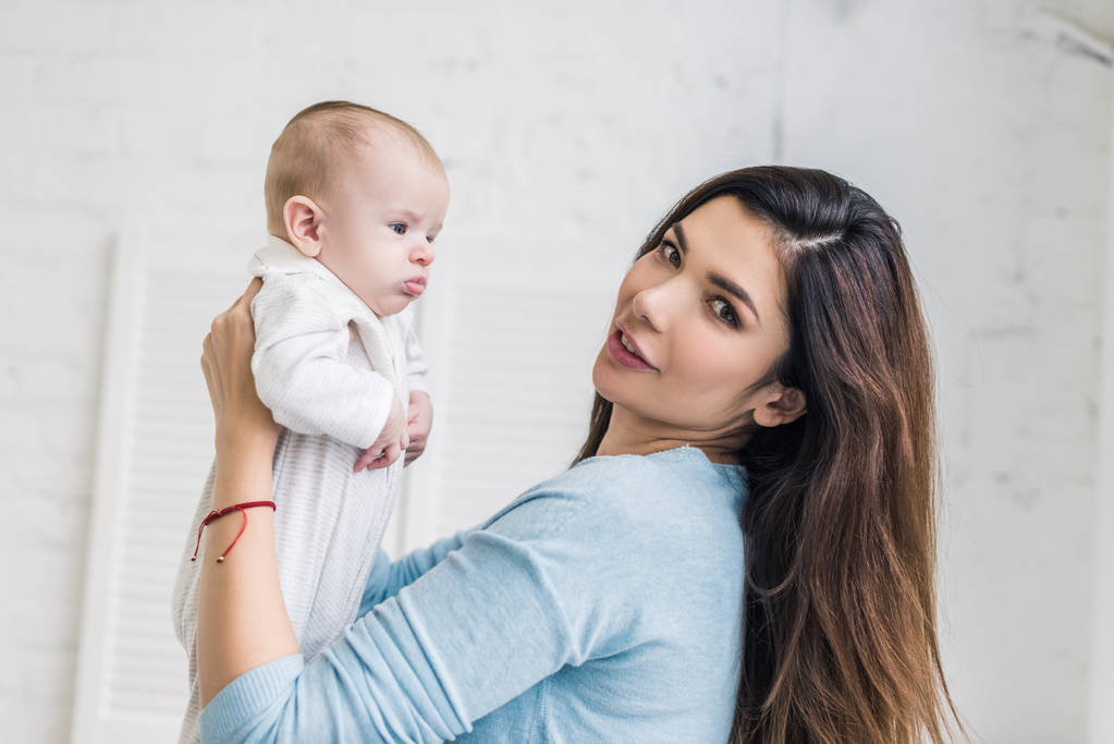 vista lateral de la joven madre mirando a la cámara mientras sostiene al bebé en las manos
 - Foto, Imagen