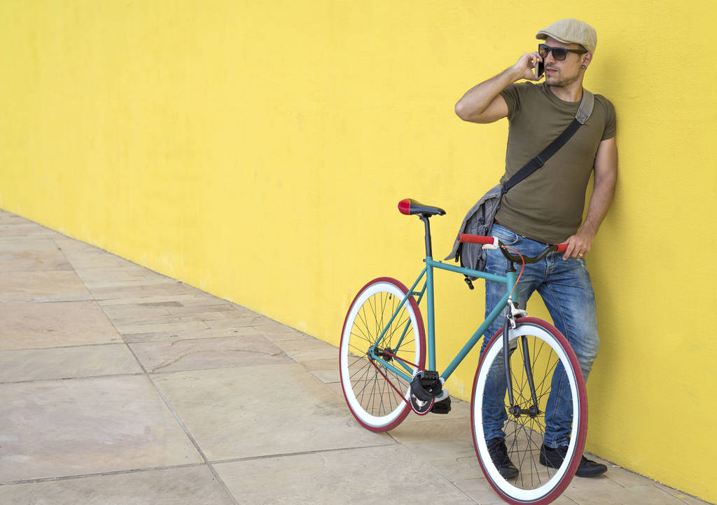 Mladý muž mluví na smartphonu stoje na kole žluté zdi na chodníku v městě - Fotografie, Obrázek