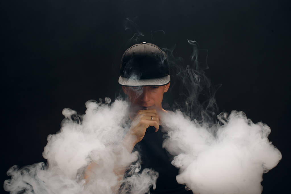 Vaporizzatore. Ritratto di un bel ragazzo bianco in un moderno berretto nero che vaporizza e sfoggia sbuffi di vapore da una sigaretta elettronica
 - Foto, immagini