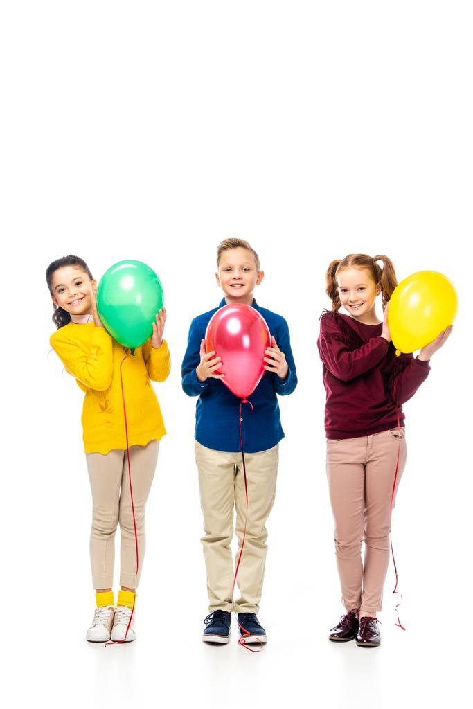 χαριτωμένο αγόρι και μαθήτριες χαμογελώντας, κρατώντας πολύχρωμα μπαλόνια και βλέπουν τα φωτογραφικών μηχανών που απομονώνονται σε λευκό - Φωτογραφία, εικόνα