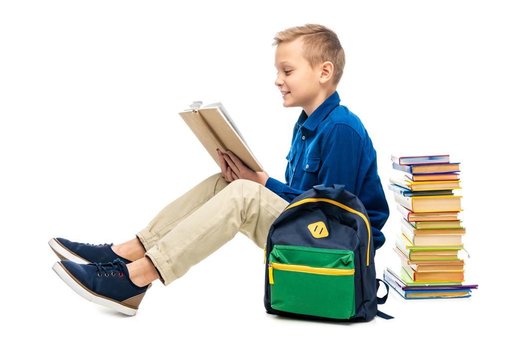 smiling boy reading book and sitting near backpack isolated on white - Photo, Image
