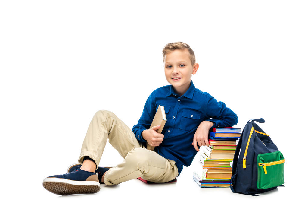 Улыбающийся школьник держит книгу, смотрит в камеру и сидит рядом с рюкзаком и стопкой книг изолированных на белом
 - Фото, изображение