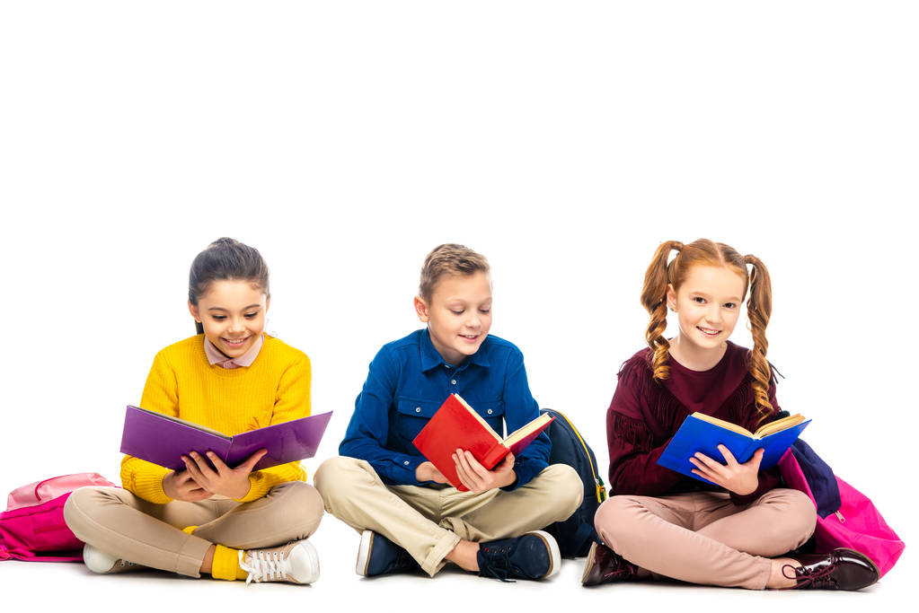 Улыбающиеся школьники сидят и читают книги с разноцветными обложками, изолированными на белом
 - Фото, изображение