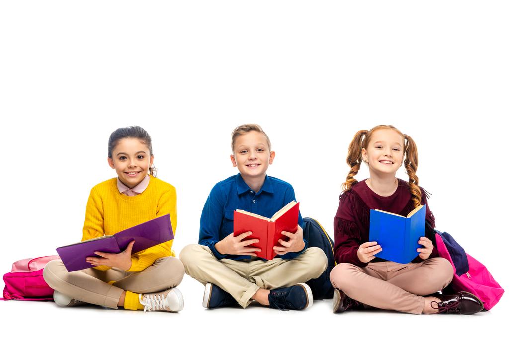 escolares alegres sentados, sosteniendo libros y mirando a la cámara aislada en blanco
 - Foto, imagen