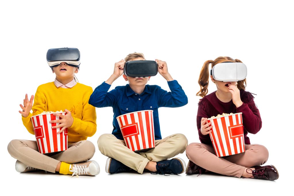 verblüffte Kinder mit Virtual-Reality-Headsets auf Köpfen, die gestreifte Pappeimer in der Hand halten und Popcorn isoliert auf weiß essen - Foto, Bild