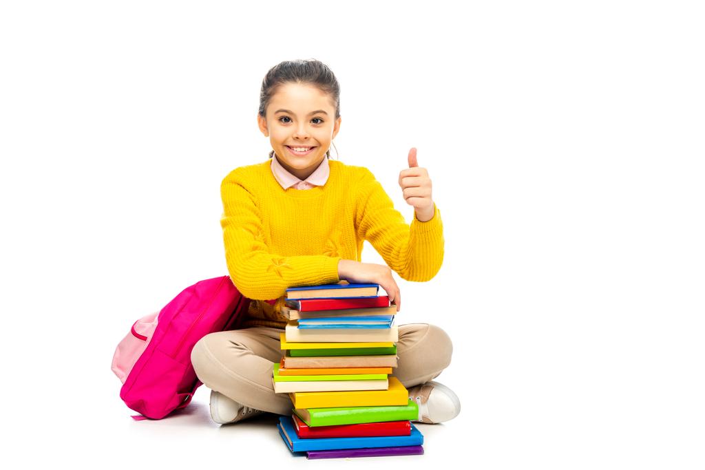 smiling schoolgirl sitting near stack of books and pink backpack, looking at camera and showing thumb up isolated on white - Photo, Image