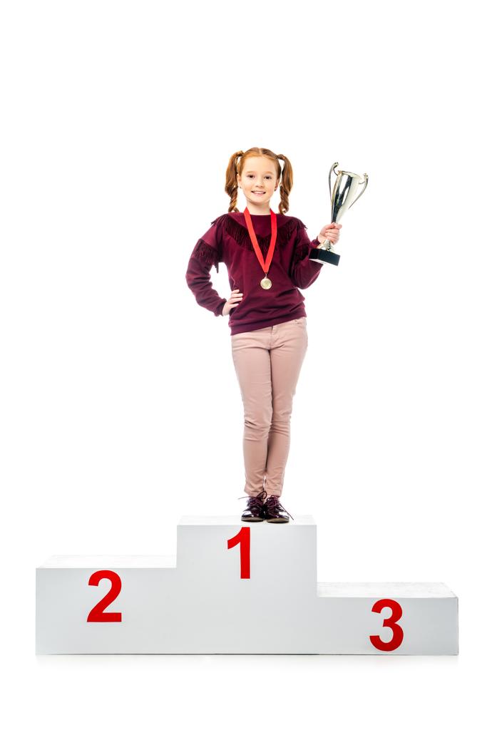 glimlachend schoolmeisje met medaille permanent op het podium van de winnaar, het bezit van trofee cup en het kijken naar camera geïsoleerd op wit - Foto, afbeelding