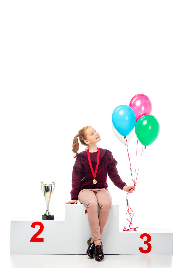 lachende schoolmeisje zittend op winnaar podium in de buurt van trofee cup en houdt van kleurrijke ballonnen geïsoleerd op wit - Foto, afbeelding