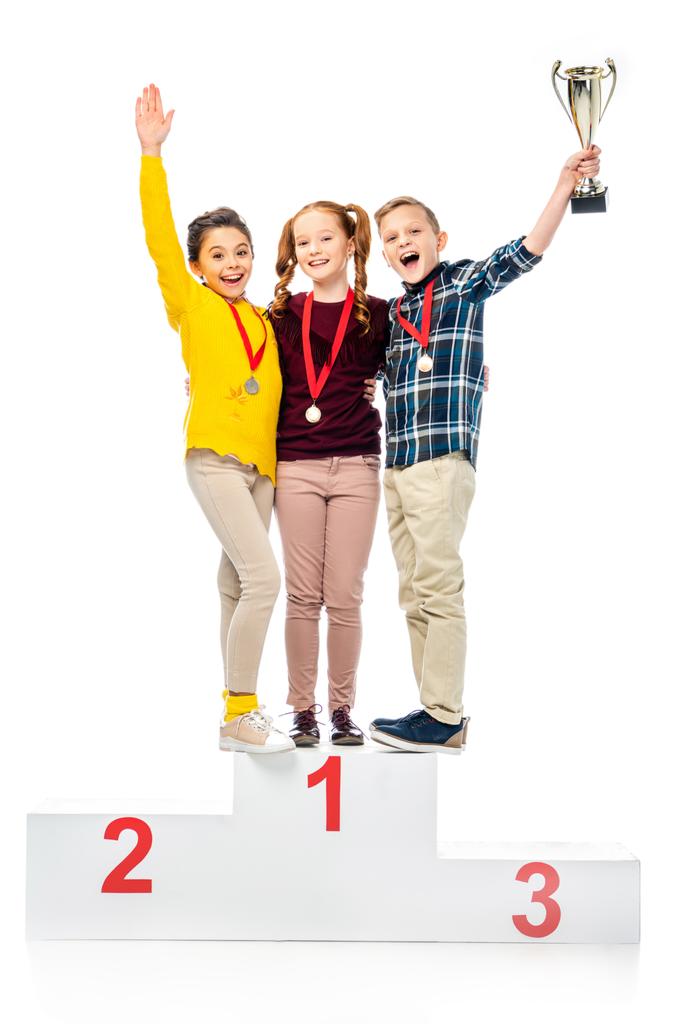 happy kids with medals and trophy cup standing on winner pedestal, shouting and looking at camera isolated on white - Photo, Image