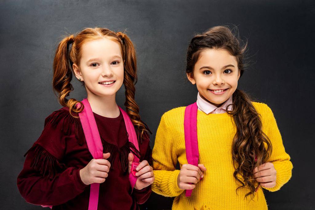 усміхнені школярки з рожевими рюкзаками дивиться на камеру на чорному тлі
 - Фото, зображення