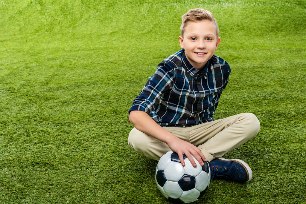 少年サッカー ボールの手で芝生の上に座って カメラ目線の笑顔 ロイヤリティフリー写真 画像素材