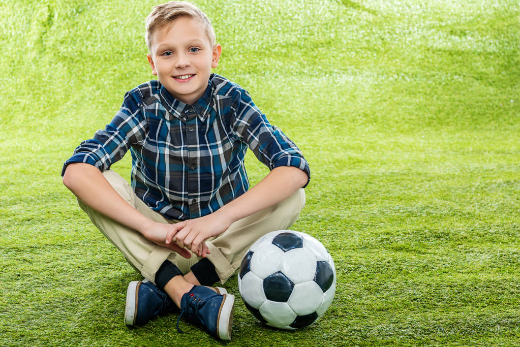 усміхнений хлопчик сидить на газоні біля футбольного м'яча і дивиться на камеру
 - Фото, зображення