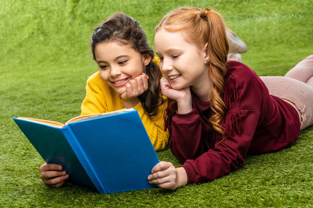 smiling schoolgirls lying on lawn and reading book - Photo, Image