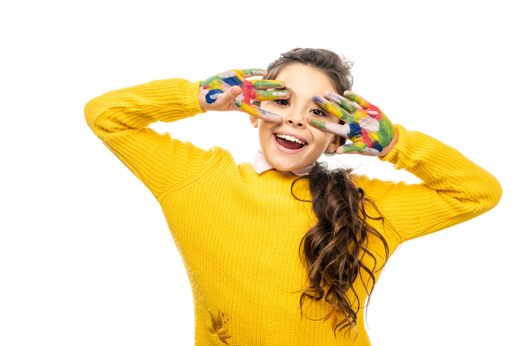 fröhliches Schulmädchen mit geöffnetem Mund im gelben Pullover, das in die Kamera blickt und Hände zeigt, die in bunten Farben isoliert auf weiß gemalt sind - Foto, Bild