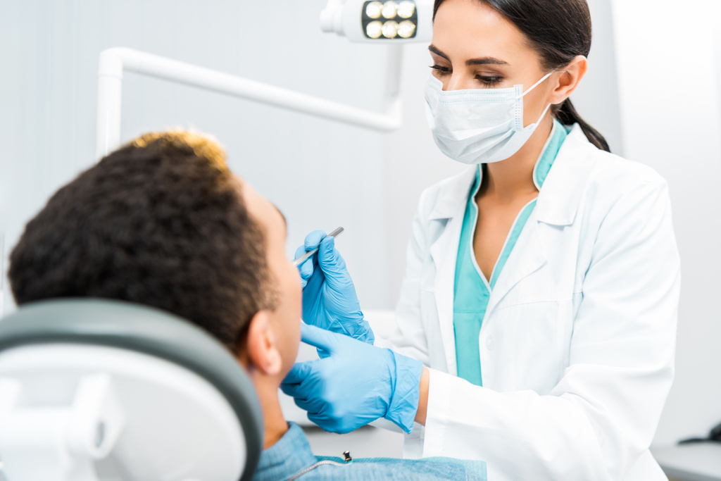 vrouwelijk tandarts in masker examencommissie Afro-Amerikaanse patiënt  - Foto, afbeelding