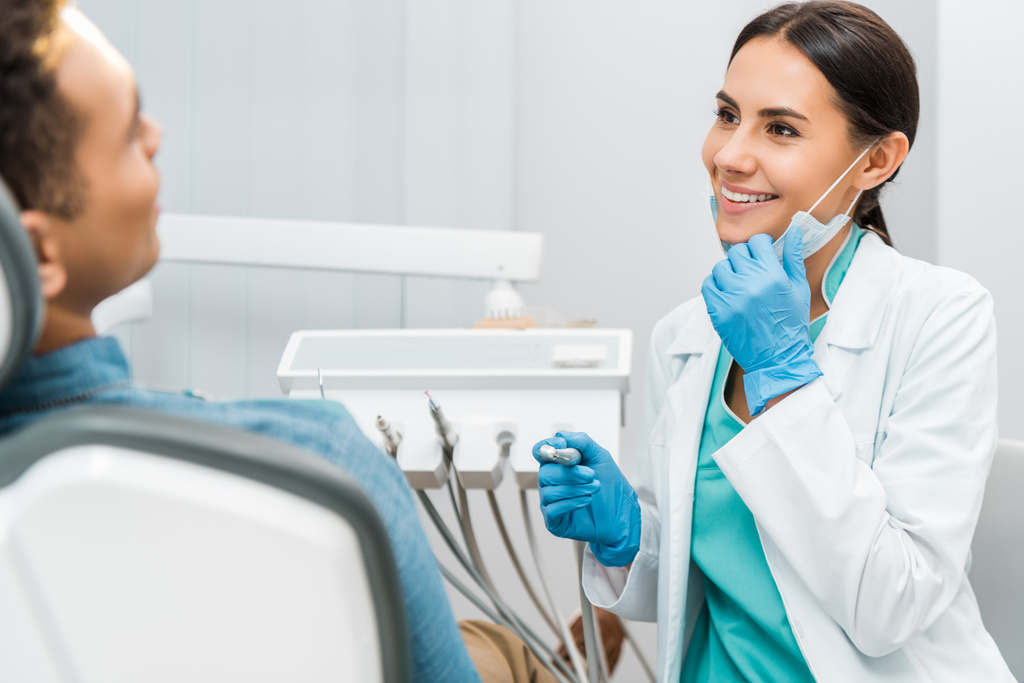 gaie dentiste femme tenant masque et foret et souriant près du patient
  - Photo, image