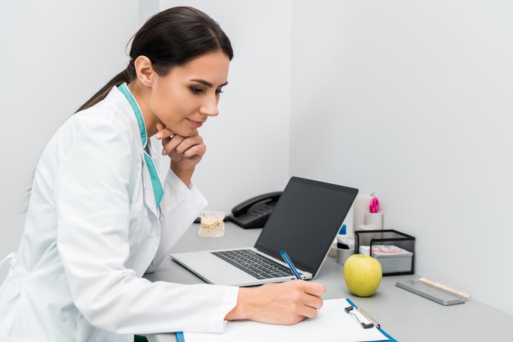 médico femenino reflexivo haciendo notas cerca de la computadora portátil
  - Foto, Imagen