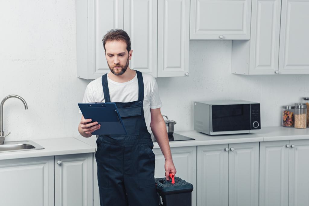 ernsthafter bärtiger Reparateur im Arbeitsalltag mit Werkzeugkiste und Blick auf Klemmbrett in der Küche zu Hause - Foto, Bild