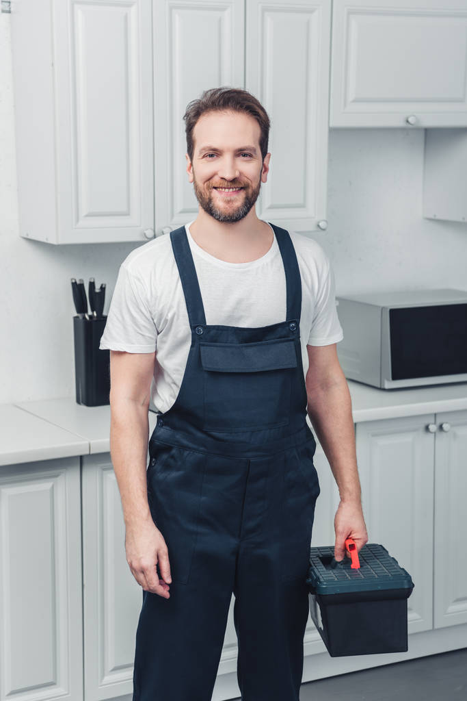 ενηλίκων χαμογελώντας γενειοφόρος επισκευαστής εργάζονται συνολικά κρατώντας εργαλειοθήκη στην κουζίνα στο σπίτι - Φωτογραφία, εικόνα