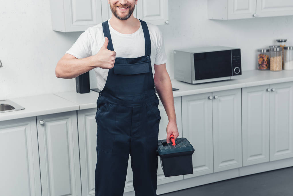 Μερική άποψη της γενειοφόρος επισκευαστής εργάζονται συνολικά κρατώντας εργαλειοθήκη και δείχνει τον αντίχειρα στην κουζίνα στο σπίτι - Φωτογραφία, εικόνα