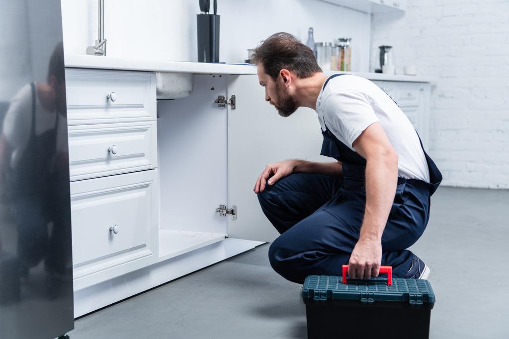 side view of adult bearded repairman with toolbox checking sink in kitchen - Photo, Image