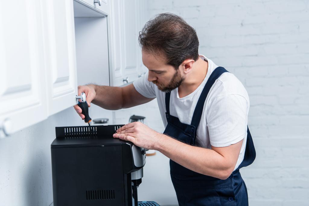 bearded handyman in working overall repairing coffee machine by screwdriver in kitchen - Photo, Image