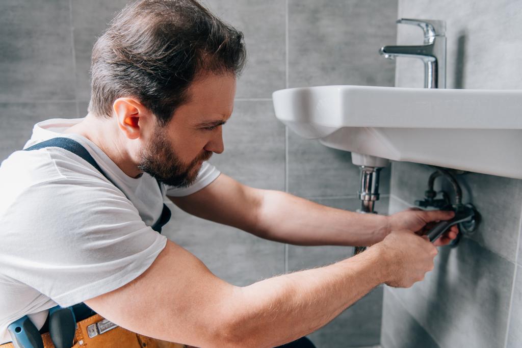adult male plumber in working overall fixing sink in bathroom  - Photo, Image