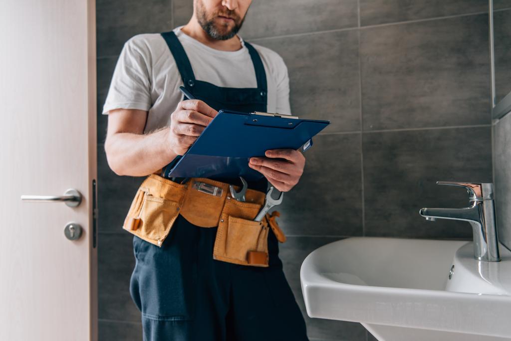 Teilansicht eines männlichen Klempners mit Werkzeuggürtel-Schrift in Klemmbrett nahe kaputtem Waschbecken im Badezimmer - Foto, Bild