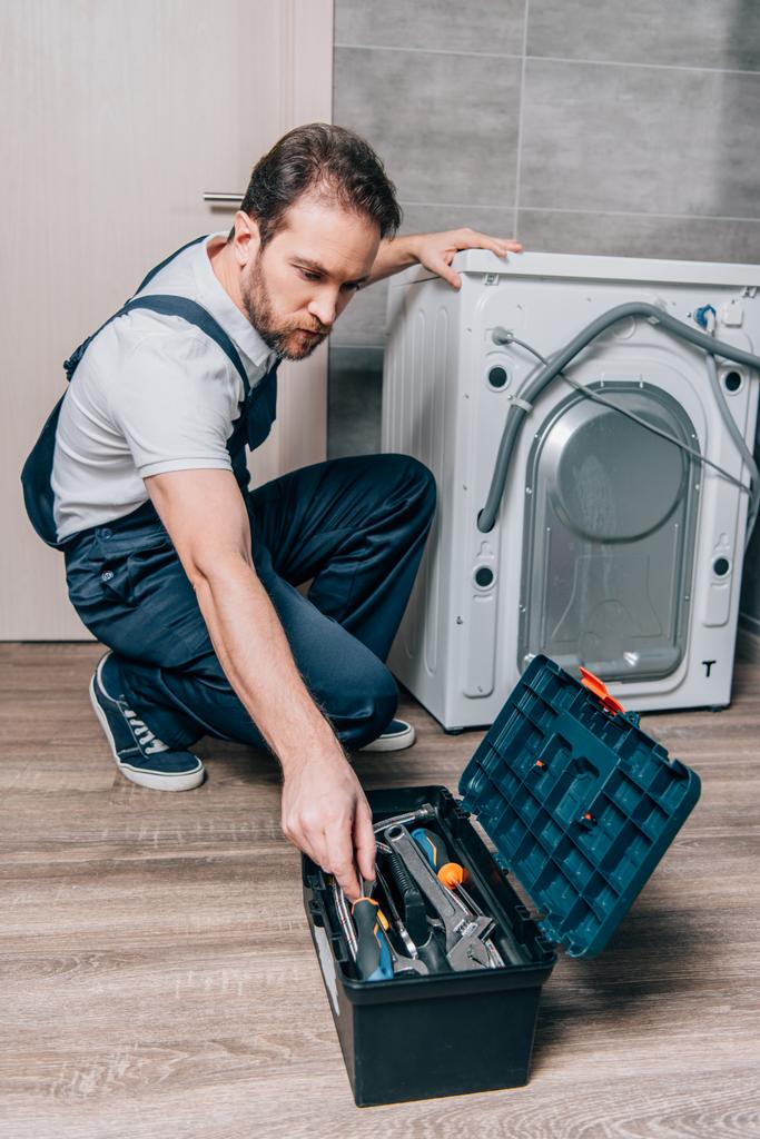 ремесленник берет инструменты из ящика для инструментов во время ремонта стиральной машины в ванной комнате
 - Фото, изображение