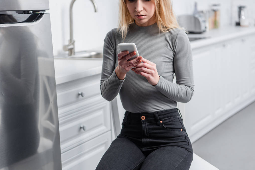 Μερική άποψη του ξανθού κοριτσιού χρησιμοποιώντας smartphone στην κουζίνα  - Φωτογραφία, εικόνα