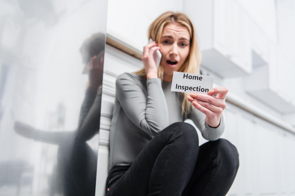 shocked young woman holding card with lettering home inspection and talking on smartphone near broken refrigerator in kitchen  - Photo, Image
